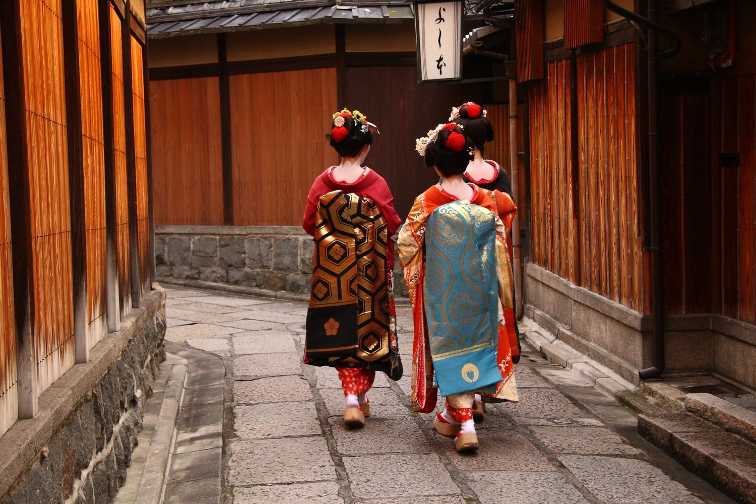 Two geiko in formal kimono walking a lantern-lined lane in Gion, Kyoto's historic geisha district