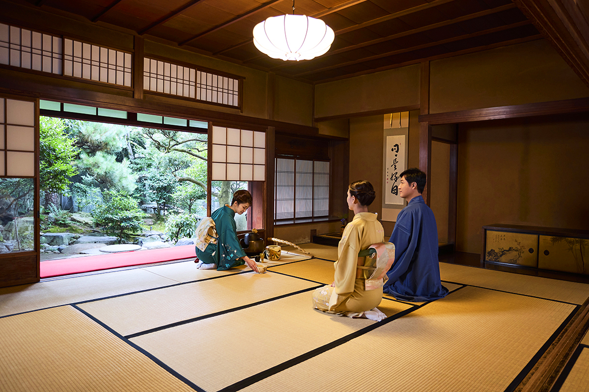 Dos invitados sentados en kimono observan a una mujer arrodillada, de kimono verde, preparar el té sobre tatami en una sala japonesa revestida de madera que se abre a un jardín.