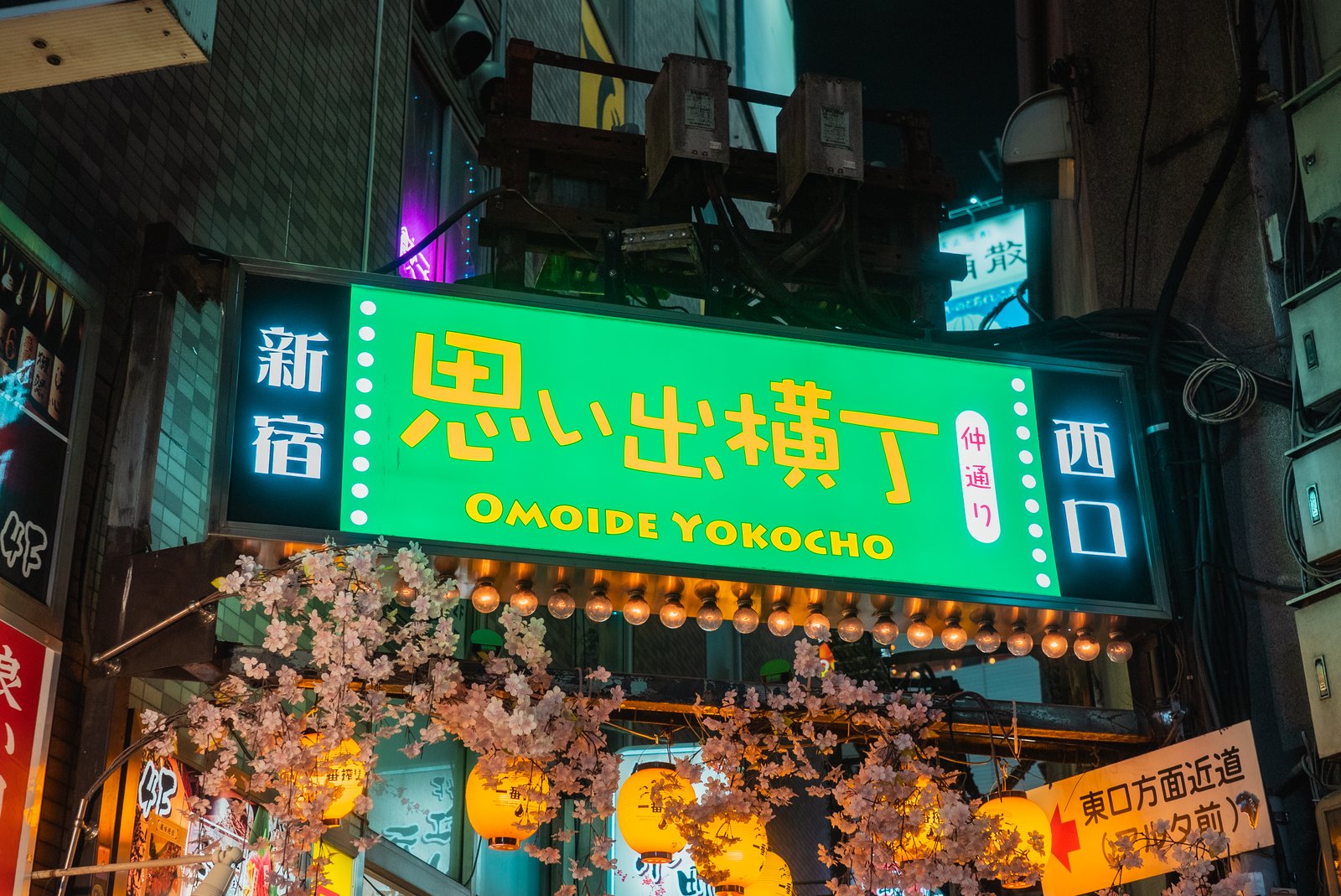 Un letrero rectangular verde iluminado que dice 'Omoide Yokocho' en japonés e inglés cuelga sobre la entrada de un callejón estrecho, enmarcado por guirnaldas de bombillas redondas y cerezos en flor, con farolillos de papel brillando debajo.