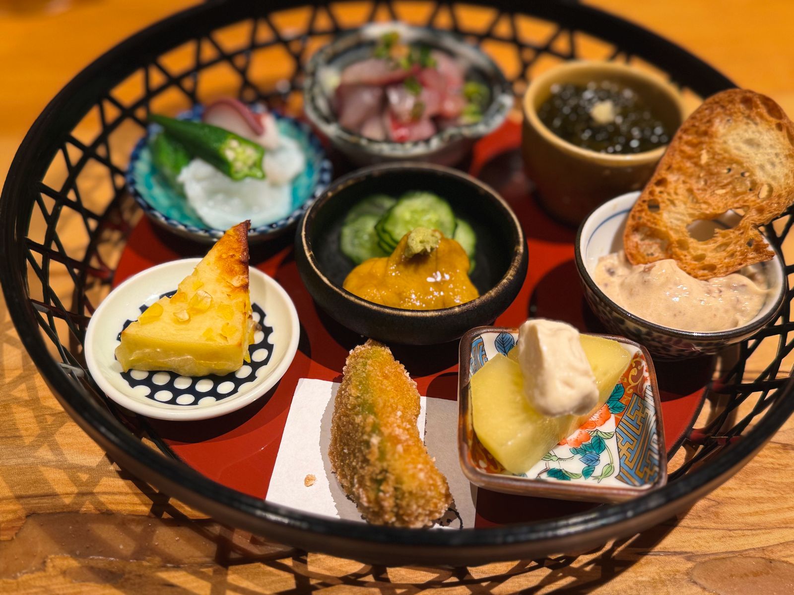 An assortment of seasonal Japanese appetisers — sashimi, tempura, tamagoyaki, pickles and more — arranged on a red tray, shot from above